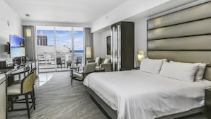 A hotel room with a large bed, a desk with a computer, a chair, and a TV. There is a view of the city through large windows.