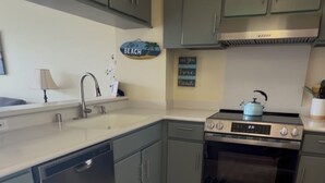 A kitchen with grey cabinets, a stainless steel oven, a dishwasher, and a striped rug. - Panoramic Ocean/Mountain View Apartment  (Mākaha)