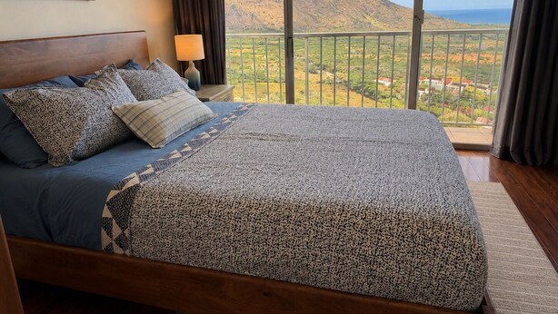 A bedroom with a large bed, a nightstand, a lamp, and a view of a mountainous landscape through a balcony. - Panoramic Ocean/Mountain View Apartment  (Mākaha)