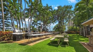 An outdoor patio with grilling stations and picnic tables, surrounded by palm trees and a well-maintained lawn. - Panoramic Ocean/Mountain View Apartment  (Mākaha)