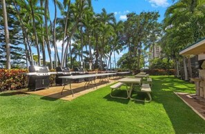 An outdoor patio with grilling stations and picnic tables, surrounded by palm trees and a well-maintained lawn. - Panoramic Ocean/Mountain View Apartment  (Mākaha)