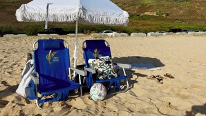 Two beach chairs under a white umbrella on a sandy beach with a hilly background. - Panoramic Ocean/Mountain View Apartment  (Mākaha)