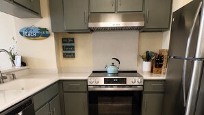 A kitchen with grey cabinets, a stainless steel refrigerator, a built-in oven, and a tiled floor. - Panoramic Ocean/Mountain View Apartment  (Mākaha)