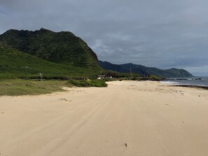 A beach with a dirt path leading to a building, surrounded by green hills and the ocean. - Panoramic Ocean/Mountain View Apartment  (Mākaha)