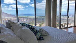A bedroom with a large bed, white bedding, and a view of the ocean and cityscape through large windows. - Panoramic Ocean/Mountain View Apartment  (Mākaha)