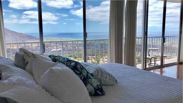 A bedroom with a large bed, white bedding, and a view of the ocean and cityscape through large windows. - Panoramic Ocean/Mountain View Apartment  (Mākaha)