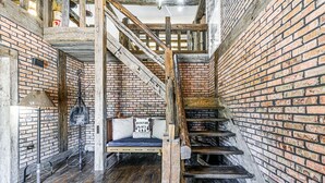 Una escalera rústica con barandillas de madera y un muro de ladrillo. Se ve una cama con un almohadón y un letrero que dice 'Adventure Awaits'.