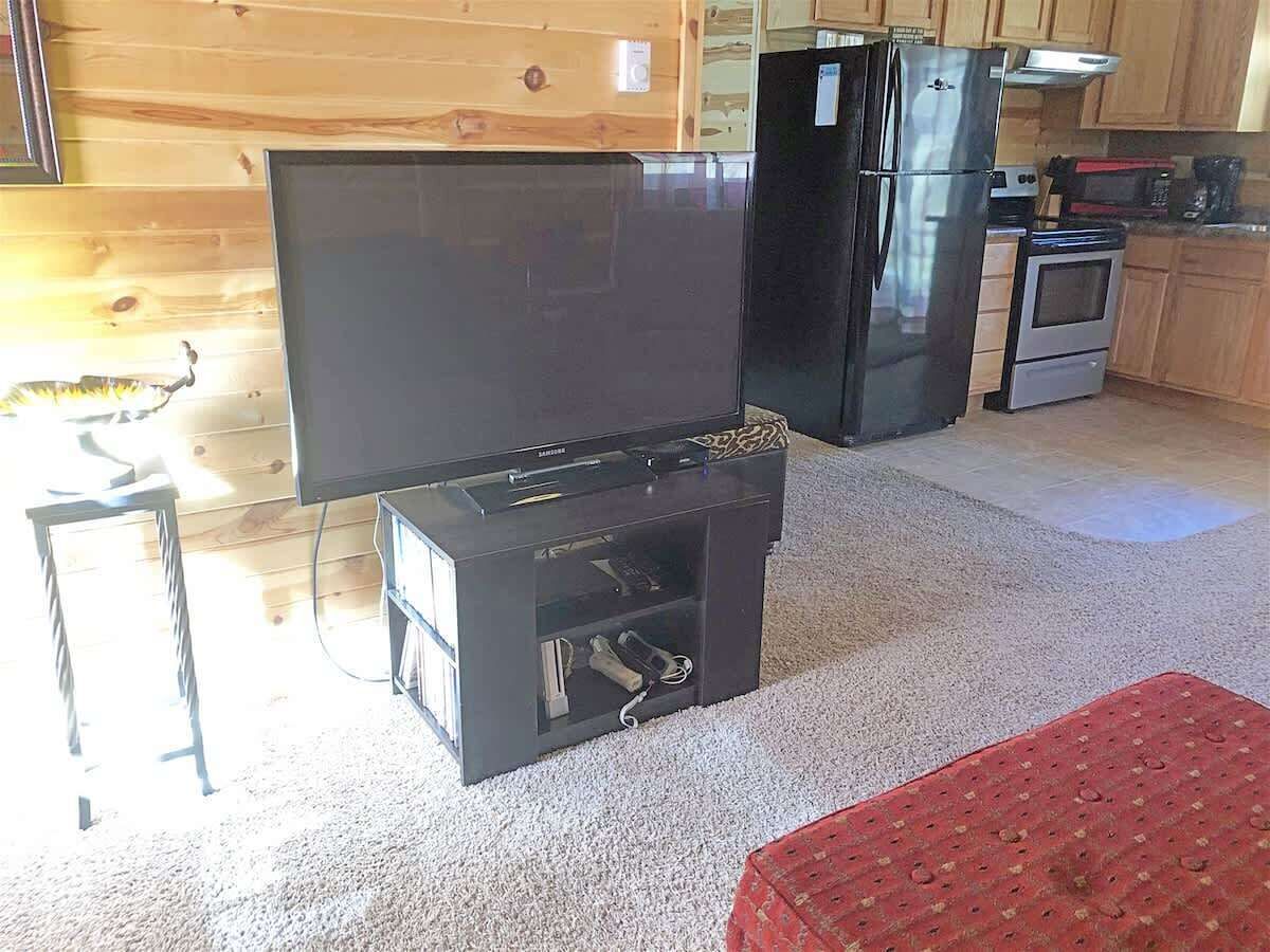 A living room with a flat-screen TV, a black entertainment unit, a refrigerator, and a kitchen area with a microwave and oven.