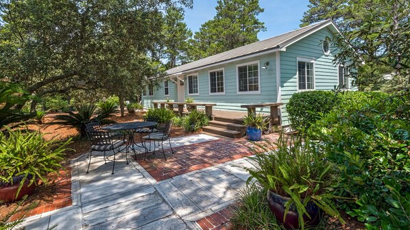 A light blue house with a patio, surrounded by lush greenery and a brick pathway.