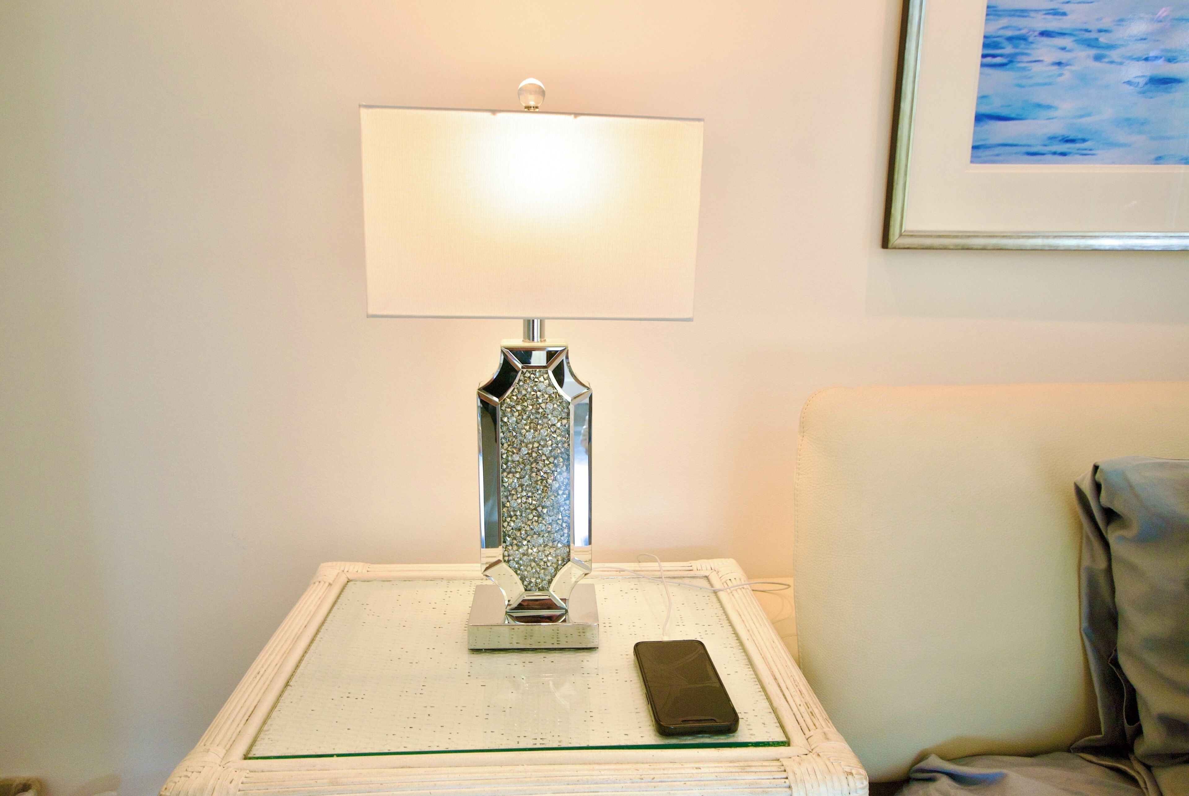 Une table de chevet avec une lampe, un téléphone intelligent et un tableau encadré accroché au mur.