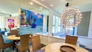 Un comedor moderno con sillas de mimbre, una mesa redonda y un espejo grande que refleja un mural tropical.
