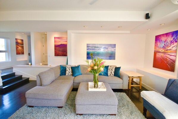 Ein modernes Wohnzimmer mit einem grauen Ecksofa, einem kleinen Tisch und einer Vase mit Blumen. Eine Treppe führt zu einer oberen Ebene, und die Wände sind mit Kunstwerken geschmückt.