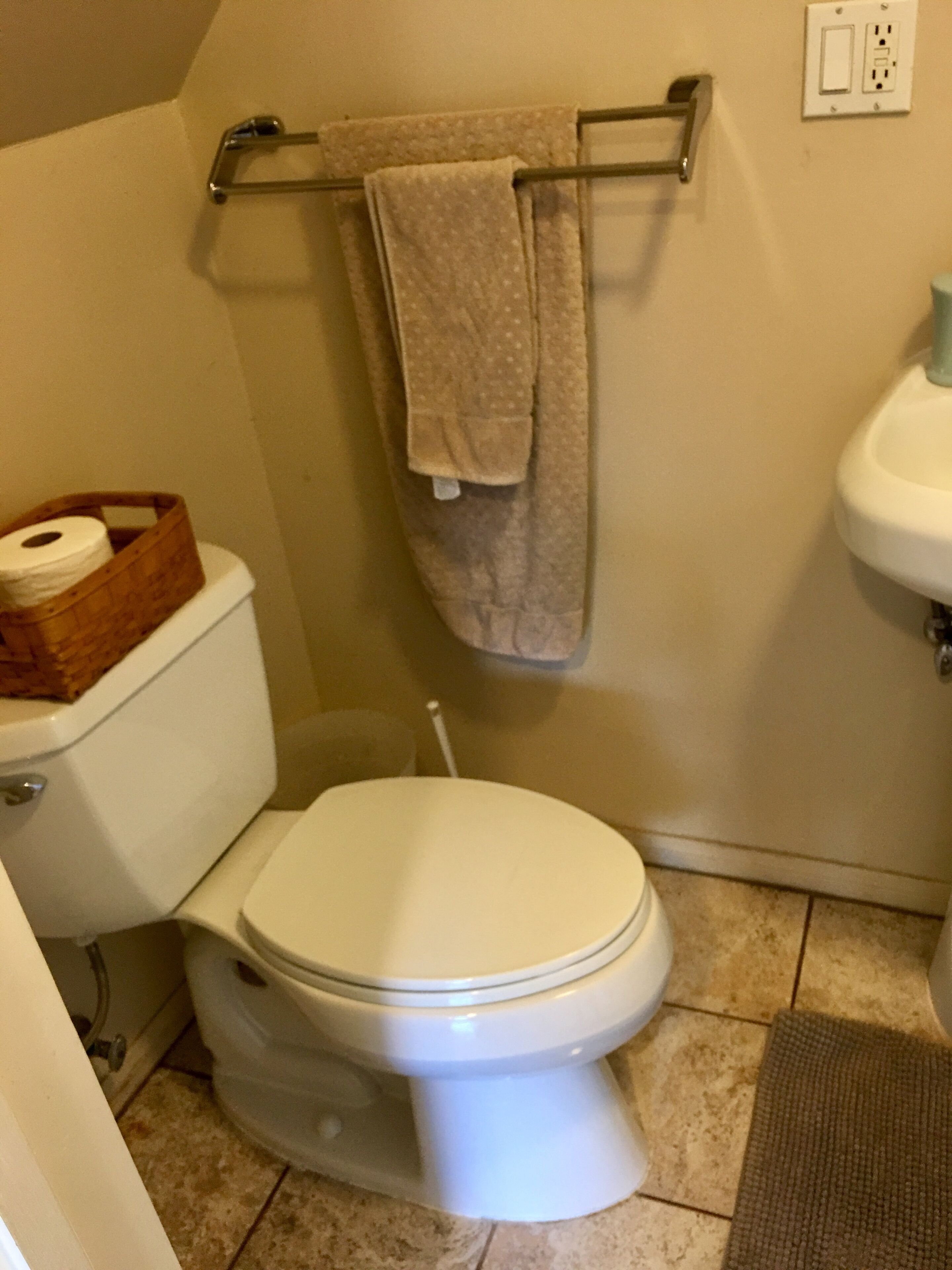A bathroom with a toilet, a towel rack with a towel, a wooden basket with toilet paper, and a sink.