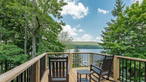 Eine Holzterrasse mit zwei Schaukelstühlen und einem Tisch, mit Blick auf einen See und bewaldete Hügel.