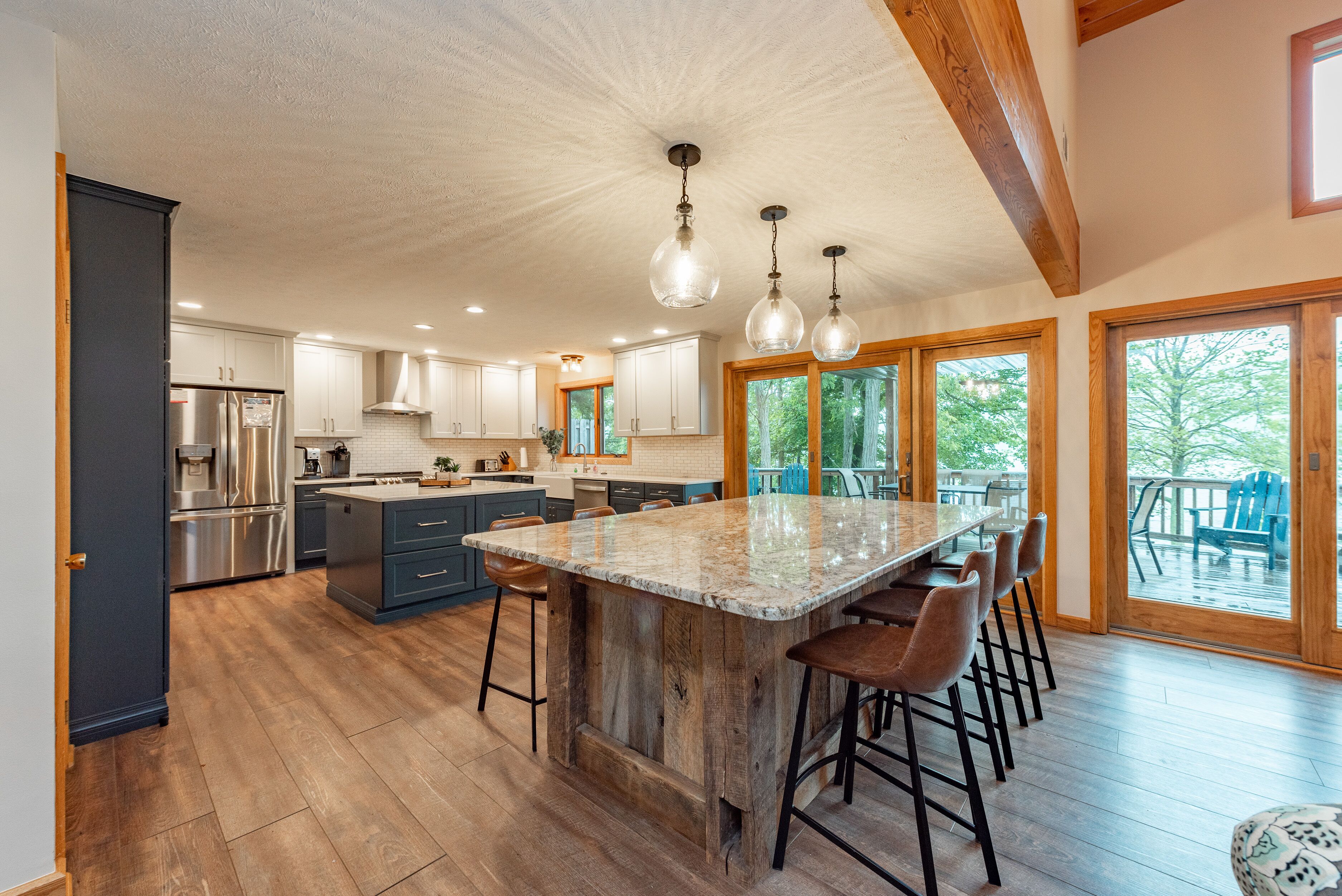 A spacious kitchen with a large island, wooden ceiling beams, and a view of the outdoors through large windows.