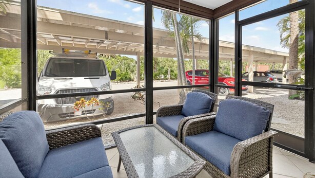 Un área de estar exterior techada con mobiliario de mimbre y una mesa de vidrio, con vista a un estacionamiento con vehículos.