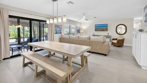 Un'ampia zona giorno con un tavolo da pranzo in legno e panche, un divano e una poltrona in vimini. Si nota un'ampia finestra con persiane, una ventola a soffitto e opere d'arte alle pareti.