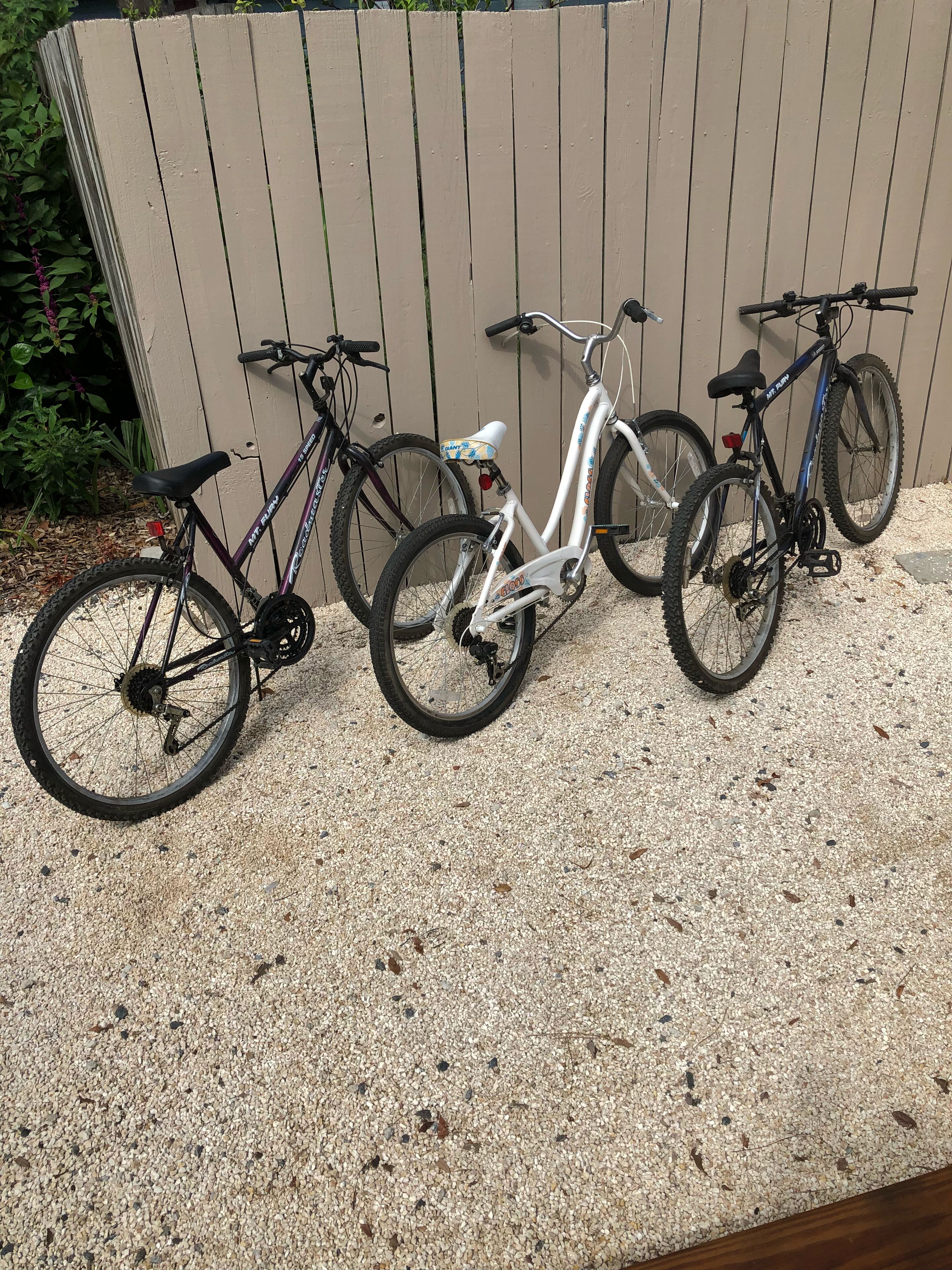 Three bicycles parked on a gravel surface against a wooden fence.