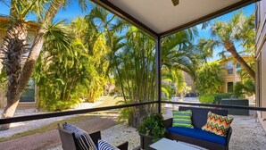 A covered outdoor seating area with wicker furniture, a small table, and a view of palm trees and a building.