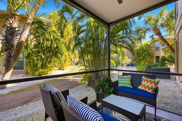 A covered outdoor seating area with wicker furniture, a small table, and a view of palm trees and a building.