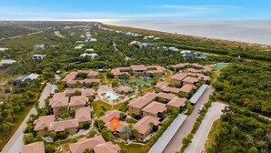 An aerial view of a resort with multiple buildings, a central swimming pool, and a beach in the background.