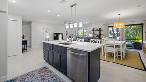 A modern kitchen with a central island, a dining area with white furniture, and a large window with a view of greenery.