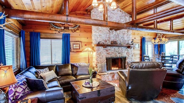 Interior acogedor de una cabaña de troncos con chimenea de piedra, muebles de cuero y techo de madera rústica.