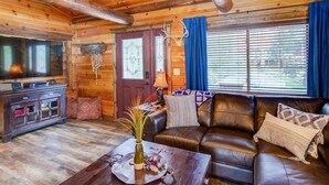 Interior acogedor de una cabaña de troncos con un sofá de cuero, un mueble de madera para entretenimiento y un amplio ventanal con cortinas.