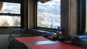 Una habitación con cama, ventana con vista a un bosque y montañas, una mesita con un tapete rojo y una silla.