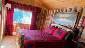 Un dormitorio de cabaña acogedor con una cama grande, cortinas de pata de gallo, una ventana con vistas y un cuadro en la pared.