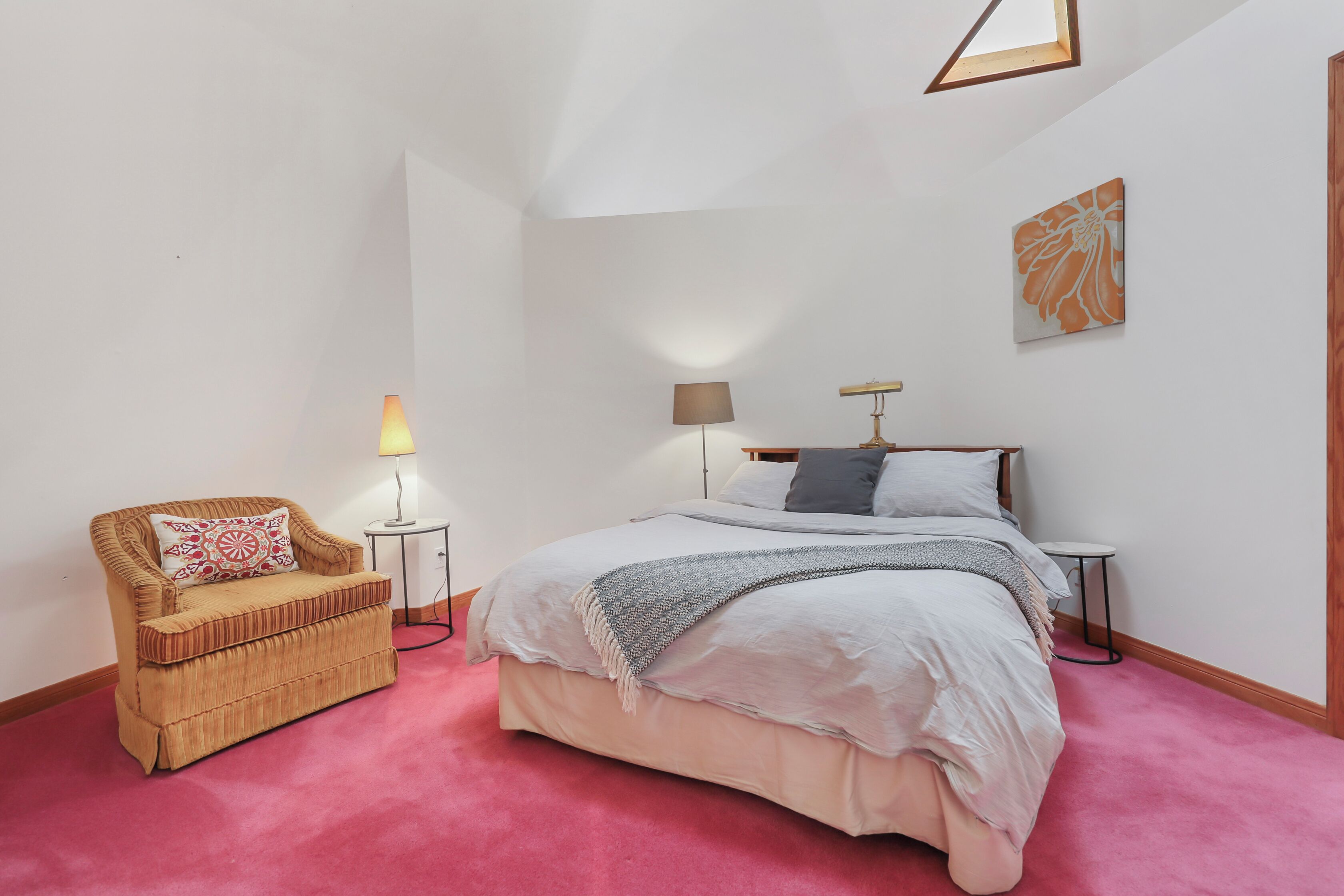 A bedroom with a bed, a chair, two bedside tables, a lamp, and a wall art piece.