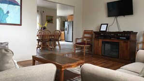 Un soggiorno con un tavolino in legno, due divani beige, una TV a parete e, sullo sfondo, una zona cucina.