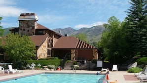 Ein Schwimmbad mit Liegestühlen, ein Steinhaus mit einem Turm und Berge im Hintergrund.