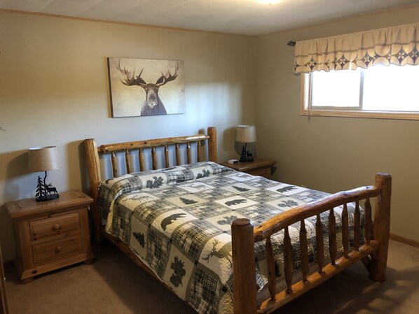 A bedroom with a wooden bed, two bedside tables, a lamp, a quilt with animal prints, a wall painting of a deer, and a window with a valance.