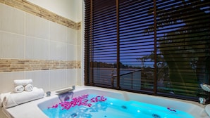 A bathroom with a large bathtub filled with water and rose petals, a towel rack with towels, and a window with blinds.