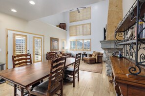 Un comedor amplio con mesa y sillas de madera, chimenea con revestimiento de piedra y ventanas grandes con contraventanas de madera.