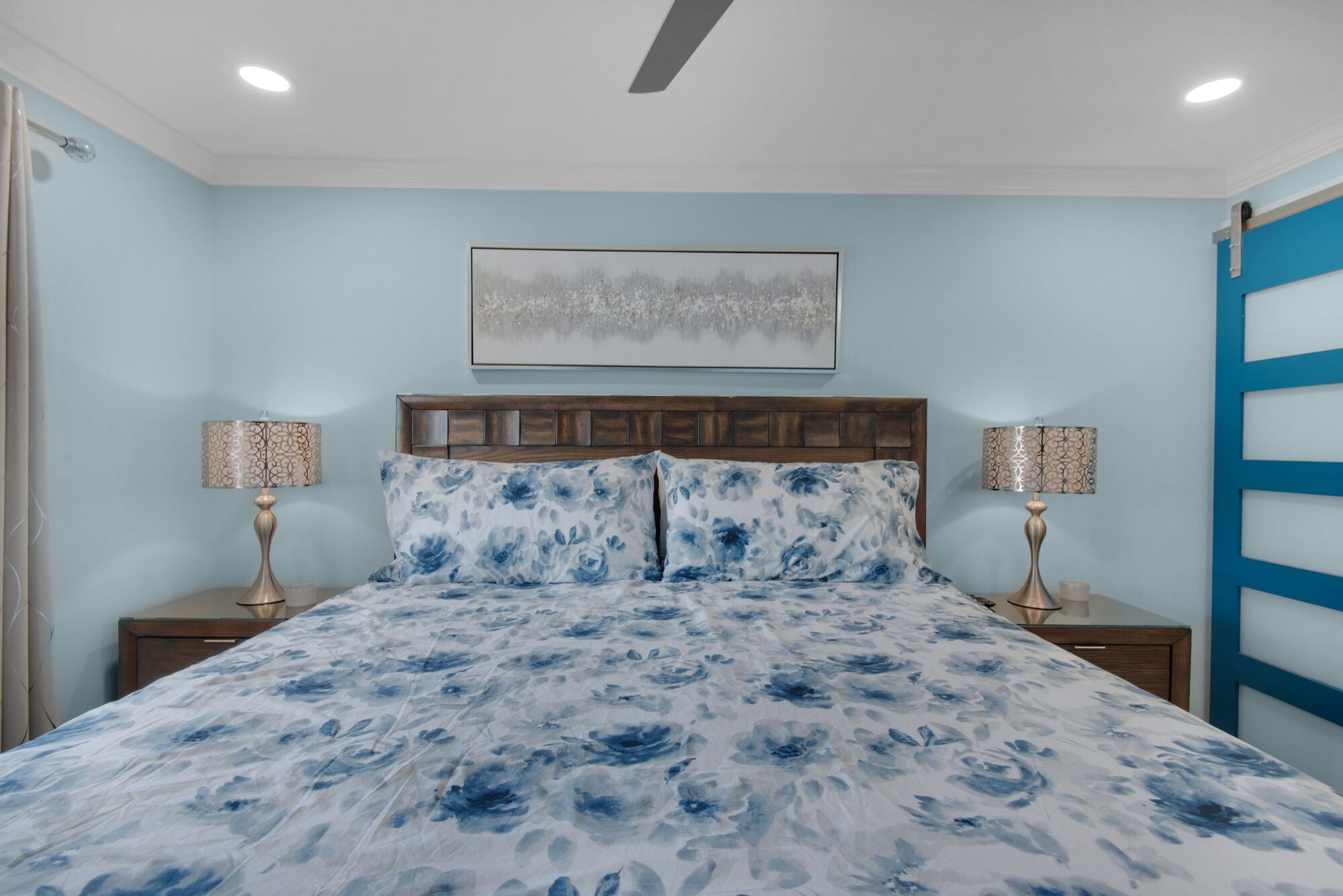 Une chambre à coucher comprenant un lit, deux tables de chevet, une tête de lit, un ventilateur de plafond et une œuvre d’art accrochée au mur.