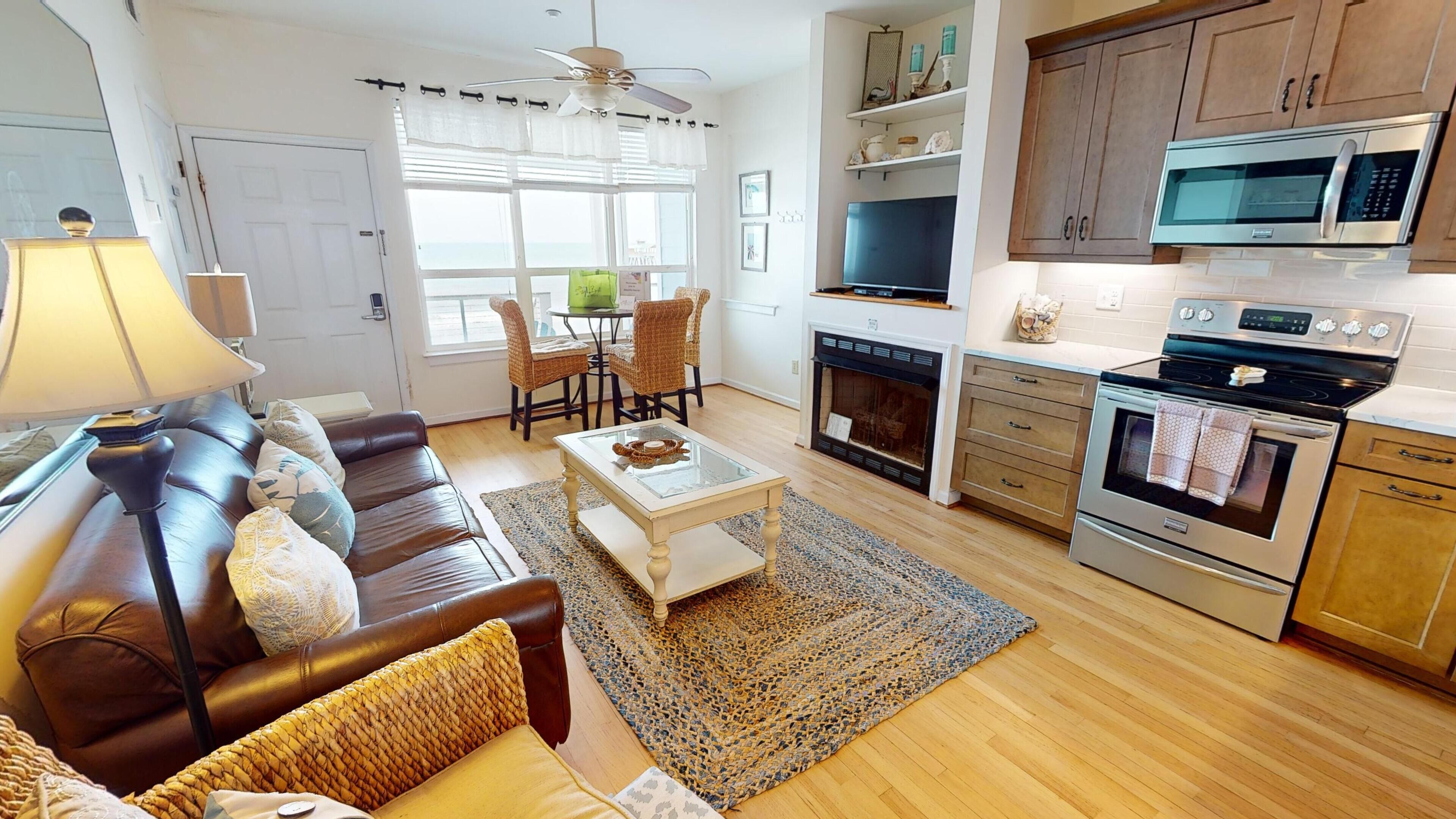 A living room with a sofa, coffee table, and a fireplace. There is a kitchen with a microwave, oven, and cabinets.