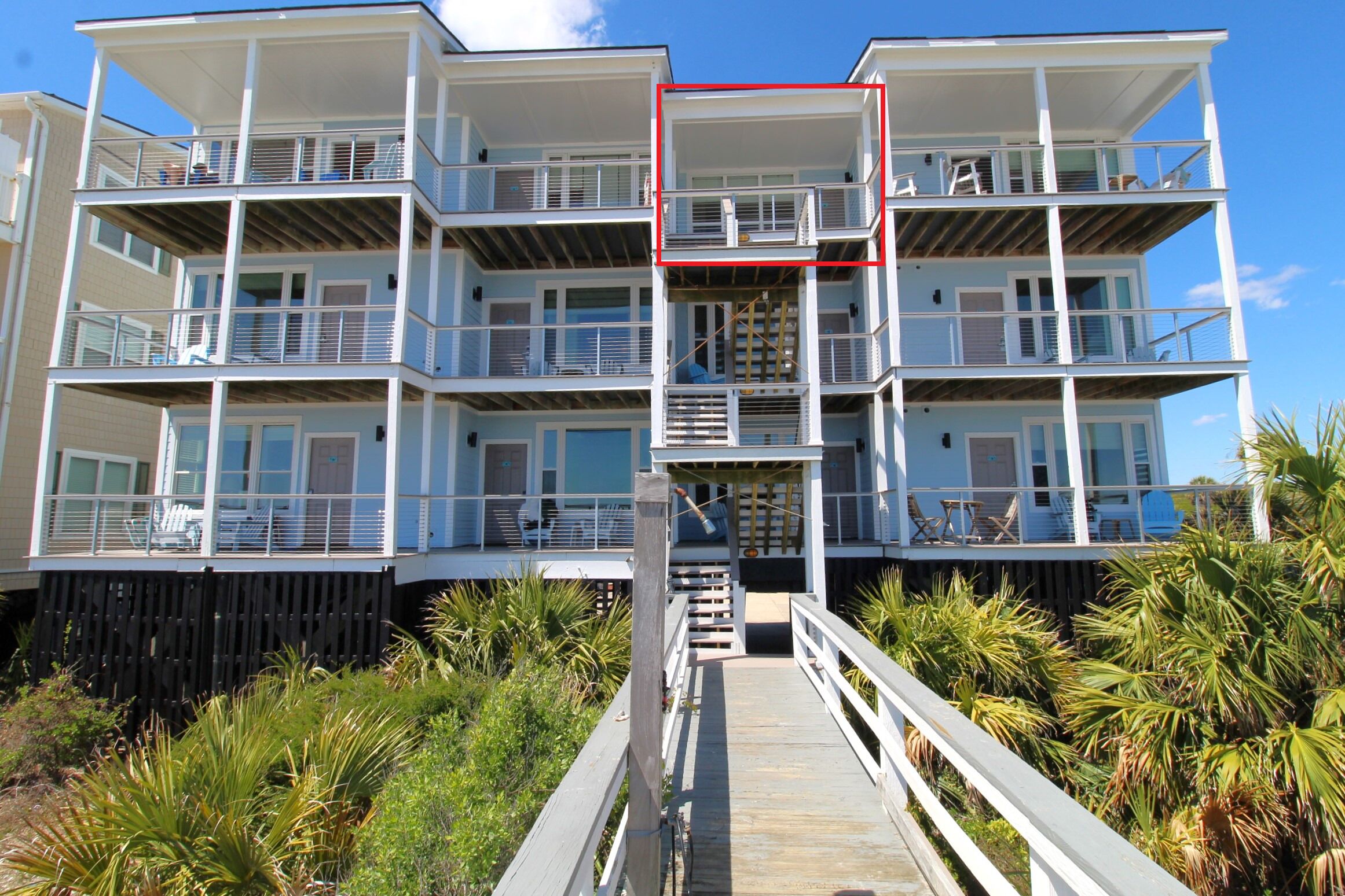 A multi-story beachfront building with balconies, a wooden deck, and surrounding vegetation.
