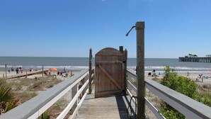 Een houten looppad dat naar een strand leidt, met een houten poort, een steiger in de verte en mensen die van het strand genieten.