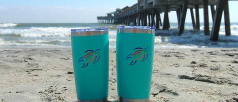 Deux gobelets turquoise ornés d’un logo coloré, posés sur une plage de sable, avec une jetée en arrière-plan.