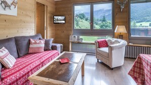 L’intérieur chaleureux d’un chalet, avec un décor en bois, un canapé, un fauteuil, une table et une grande fenêtre donnant sur un paysage montagneux.