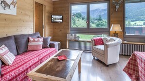 L’intérieur chaleureux d’un chalet, avec un décor en bois, un canapé, un fauteuil, une table et une grande fenêtre donnant sur un paysage montagneux.