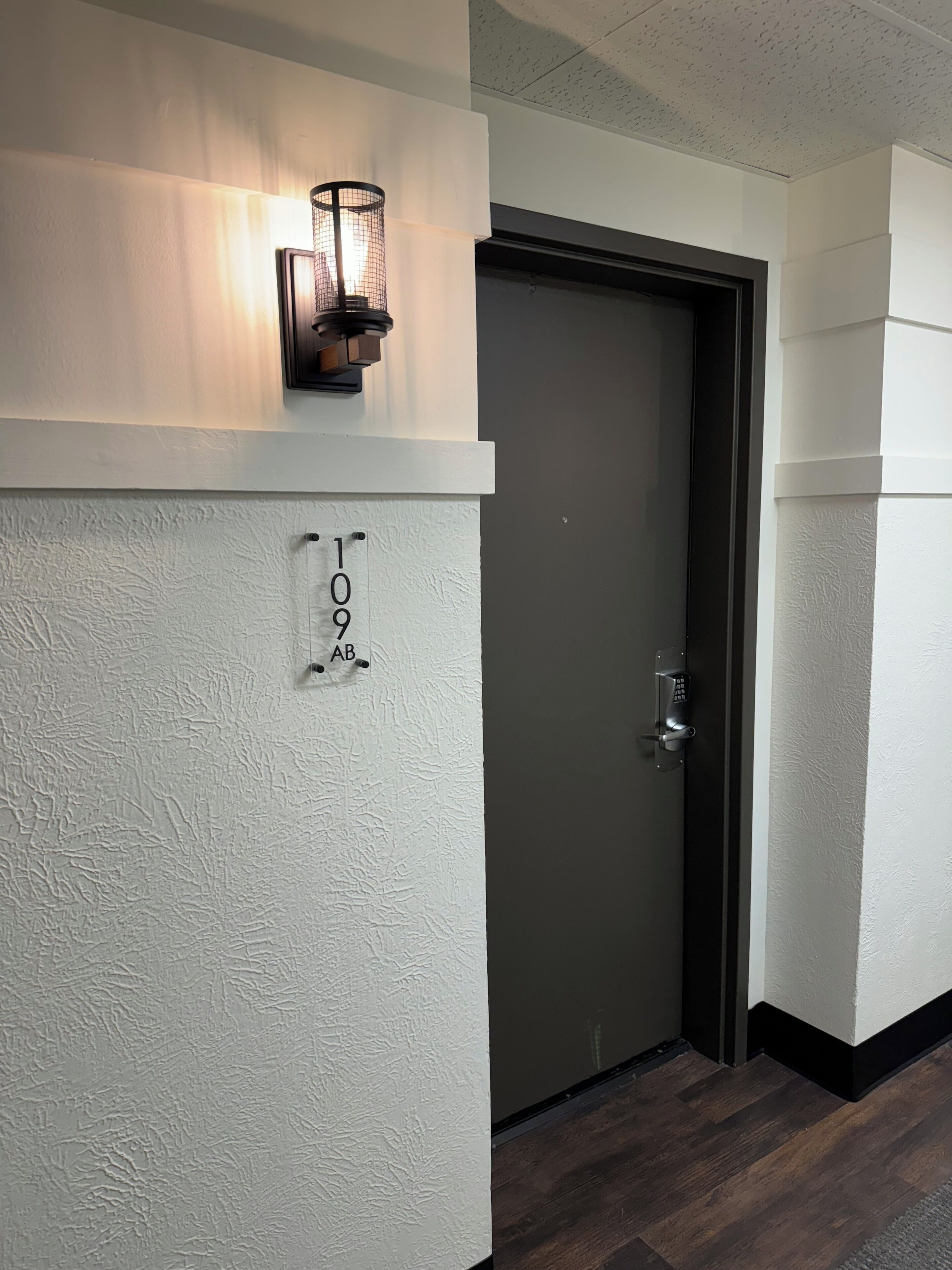 Un pasillo con pared texturizada, una puerta con cerradura electrónica y una lámpara fijada a la pared.