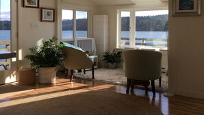 A spacious living room with a large window overlooking a body of water, a round rug, and a ceiling fan. - Secluded Overwater Eco-Cottage on Private Beach (Vashon)