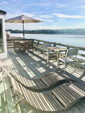 A wooden deck with outdoor seating, a table with food, and a view of a lake. - Secluded Overwater Eco-Cottage on Private Beach (Vashon)