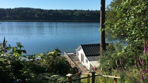 A lakeside property with a wooden deck, a white building with a dark roof, and dense greenery. - Secluded Overwater Eco-Cottage on Private Beach (Vashon)