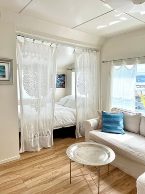 A room with a bed, a sofa, a round metal table, and a window with curtains. - Secluded Overwater Eco-Cottage on Private Beach (Vashon)