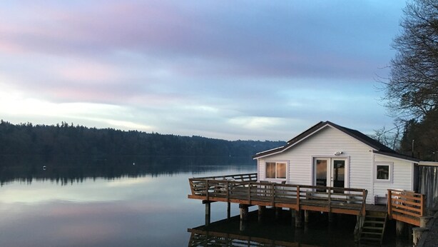 A white cabin with a wooden deck on a calm lake, surrounded by trees. - Secluded Overwater Eco-Cottage on Private Beach (Vashon)
