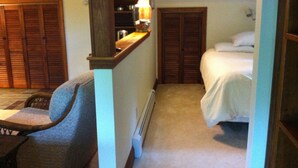 Un couloir étroit avec un lit à droite, une chaise à gauche et une lampe au-dessus d’une étagère.
